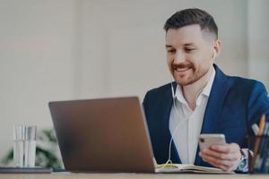 el trabajador de oficina masculino feliz utiliza poses de tecnologías modernas en el escritorio sostiene un teléfono móvil hace una videollamada a través de una computadora portátil usa auriculares vestido formalmente tiene una expresión alegre mira un seminario web de negocios foto