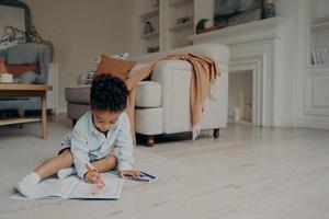 Little mixed race kid boy drawing in coloring book while spending leisure time alone at home photo