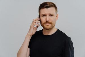 Displeased serious man has unpleasant conversation with friend holds smartphone near ear wears casual black t shirt frowns face isolated over blue background. Disappointed guy calls someone. photo