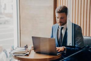 Young handsome businessman with neat beard sits by table in cafe with laptop photo