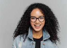 Glad woman with curly hair, wears optical glasses, denim jacket, looks straightly at camera, isolated on grey background, enjoys lovely conversation, satisfied with finished work. Facial expressions photo