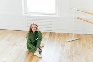 Indoor shot of satisfied good looking redhead female sits on floor, keeps legs crossed, ballet barre stands near, being professional dancer, wears sweatsuit, does stretching, poses in rehearsal room photo