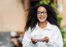 Outdoor shot of happy dark skinned lady with crisp hair, wears white jacket, drinks hot tea, has stroll across street, happy expression. Satisfied Afro woman enjoys spare time during weekend photo