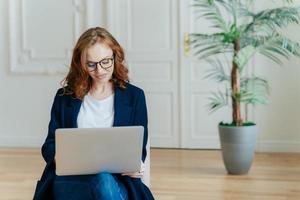 Photo of good looking ginger young woman in spectacles watches tutorial video online, poses in comfortable couch in modern apartment, dressed in elegant outfit, enjoys free internet connection
