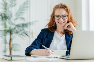 Happy successful female writes checklist in notepad, works on creative ideas for startup project, searches necessary information on laptop computer, poses at coworking space, works freelance photo