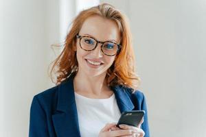 Close up shot of pretty redhead female directror has charming smile, dressed in elegant clothes, holds mobile phone, checks text message, surfs social networks, connected to wireless internet. photo