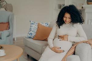 Pregnant afro american lady taking break on couch at home, talking with future baby