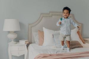 Carefree cute afro american child boy with lollipop jumping ob bed at home photo