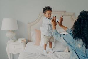 Cute happy small afro american kid playing with his mom in bedroom at home
