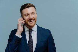 Glad male employer has telephone conversation with colleague looks away smiles gladfully wears formal suit and tie connected to roaming while having business trip poses against blue background photo