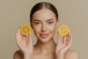 Headshot of beautiful brunette woman holds juicy lemon slices has healthy shiny skin gets vitamins from citrus stands bare shoulders indoor against beige background. Facial treatment concept