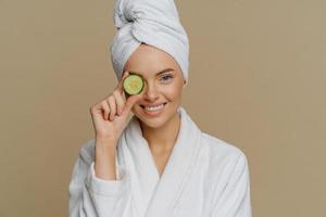 Horizontal shot of pleased refreshed woman covers eye with slice of fresh cucumber cares about skin uses organic products smiles pleasantly dressed in bath dressing gown after taking shower.
