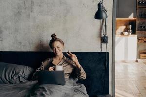encantadora jovencita alegre acostada en la cama con una taza de café y haciendo videollamadas con amigos en línea en una laptop foto