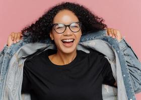 Isolated shot of curly cheerful woman tries on new denim jacket, wears optical glasses, smiles broadly, has perfect mood, poses against pink wall. Positive emotions. Afro female in stylish clothes photo