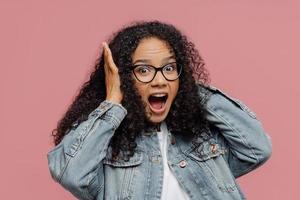 foto de una mujer afroamericana sorprendida que cubre los oídos, grita en voz alta, ignora el sonido fuerte, mantiene la boca abierta, usa anteojos y chaqueta de mezclilla, se para contra la pared rosada. omg, qué ruidoso allí