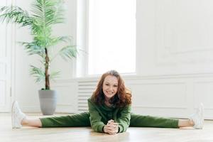 Flexible slim woman with ginger hair, does leg split, has stretching exercises, poses in home interior, has gentle smile on face, wears green tracksuit and white sneakers. Fitness model indoor photo