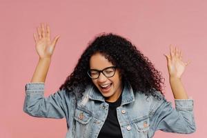 Isolated shot of beautiful happy woman with curly hairstyle, keeps hands raised, laughs from positive emotions, celebrates success has fun with friends dressed in fashionable apparel. Emotions concept photo