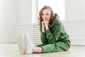 Young slim young woman keeps hands under chin, has make up, sits on floor and stretches legs, wears green sweatsuit, looks straightly at camera. People, workout and pilates exercises concept photo