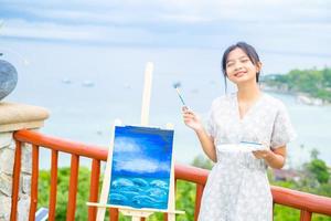 Young girl using brush drawing picture on canvas paper, at beautiful landscape view at koh tao Thailand. photo