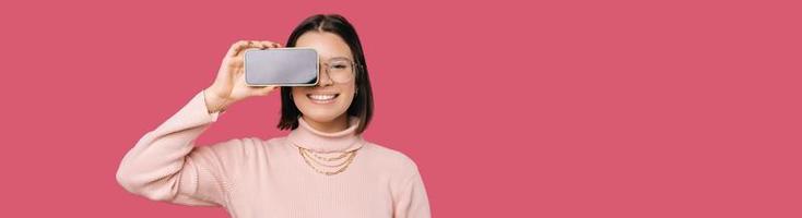 Happy young woman is hiding one eye with the phone with blank screen. photo