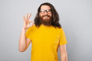 Portrait of joyful young bearded man with long hair showing ok gesture