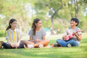 un niño tailandés-indio y una amiga tailandesa-europea se sientan y tocan el ukelele y cantan en el parque foto