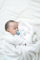 A recently born half-born baby is wrapped in a white cloth and slept in bed while sucking on a rubber nipple photo