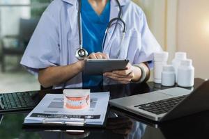 dentista concentrado sentado a la mesa con muestras de mandíbula modelo de diente y trabajando con tableta y computadora portátil en el consultorio dental clínica dental profesional. foto