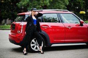 Portrait of beautiful blonde sexy fashion woman model in cap and in all black, blue corset, with bright makeup near red city car. photo
