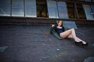 chica modelo sexy con piernas largas en traje de baño de cuerpo de lencería negra vestido combinado y chaqueta posada en el techo de un lugar industrial abandonado con ventanas. foto