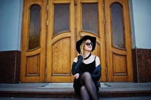 mujer rubia con vestido negro, chaqueta de cuero, gafas de sol, collares y sombrero contra puertas de madera. foto