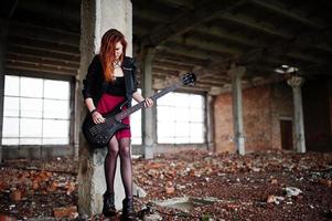 Chica punk pelirroja vestida con falda negra y roja, con bajo en un lugar abandonado. retrato de mujer gótica músico. foto