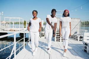 Three stylish african american friends, wear on white clothes at pier on beach. Street fashion of young black people. Black man with two african girls. photo