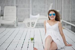 retrato de una joven fabulosa con gafas de sol y vestido sentada en un suelo de madera blanca con su bebida. foto