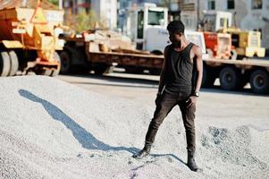 Attractive black african american guy in black muscle shirt posing on the pile of gravel in industrial zone. photo