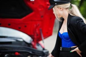 retrato de una hermosa modelo rubia de moda sexy con gorra y todo negro con maquillaje brillante cerca del coche rojo de la ciudad con el capó abierto. foto