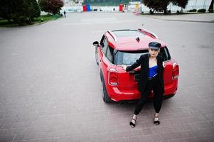 retrato de una hermosa modelo rubia de moda sexy con gorra y corsé negro y azul, con maquillaje brillante cerca del coche rojo de la ciudad. foto