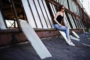 Girl at sunglasses and jeans posed at the roof of abadoned industrial place with windows. photo