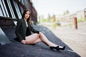 chica modelo sexy con piernas largas en traje de baño de cuerpo de lencería negra vestido combinado y chaqueta posada en el techo de un lugar industrial abandonado con ventanas. foto