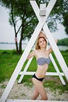 Portrait of an attractive female model posing next to the triangular white wooden construction in the park with a lake in the background. photo