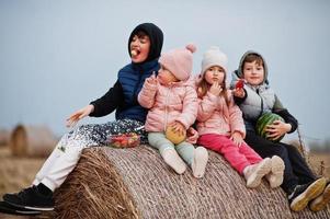 Four kids with fruits in hands sitting on haycock at field. photo