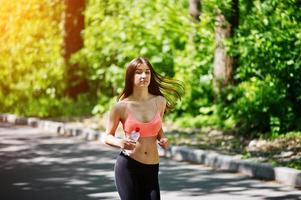 Fitness sport girl in sportswear run at road in forest with bottle water on hand, outdoor sports, urban style. photo