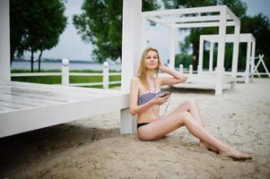 Portrait of a gorgeous young girl in bikini sitting next to the gasebo on sand and posing. photo
