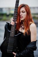 Red haired punk girl wear on black with bass guitar at the roof. photo