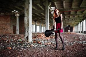 Red haired punk girl wear on black and red skirt, with bass guitar at abadoned place. Portrait of gothic woman musician. photo