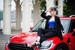 retrato de una hermosa modelo rubia de moda sexy con gorra y todo negro con maquillaje brillante sentada en el capó del coche rojo de la ciudad. foto