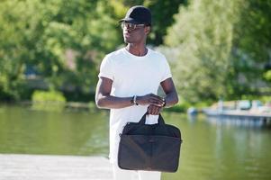 Portrait of stylish african american boy, wear on white clothes, glasses and cap with bag on hand against pier of lake. Street fashion of young black people. photo