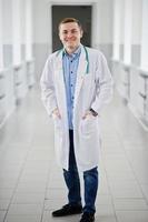 Handsome and confident young doctor in white coat with stethoscope posing in the hospital. photo