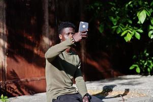 Portrait of a black african american guy sitting on the old fence and using his smartphone. photo