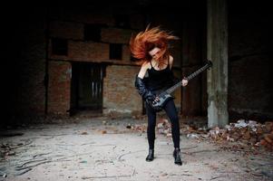 Red haired punk girl wear on black with bass guitar at abadoned place. Portrait of gothic woman musician. photo